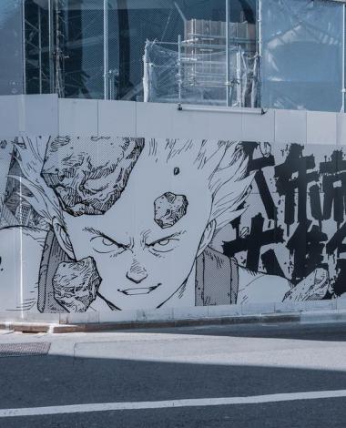 A black and white Manga mural on a white hoarding wall in what looks like an urban city street with a road in the foreground, and a glass building in the background behind the mural